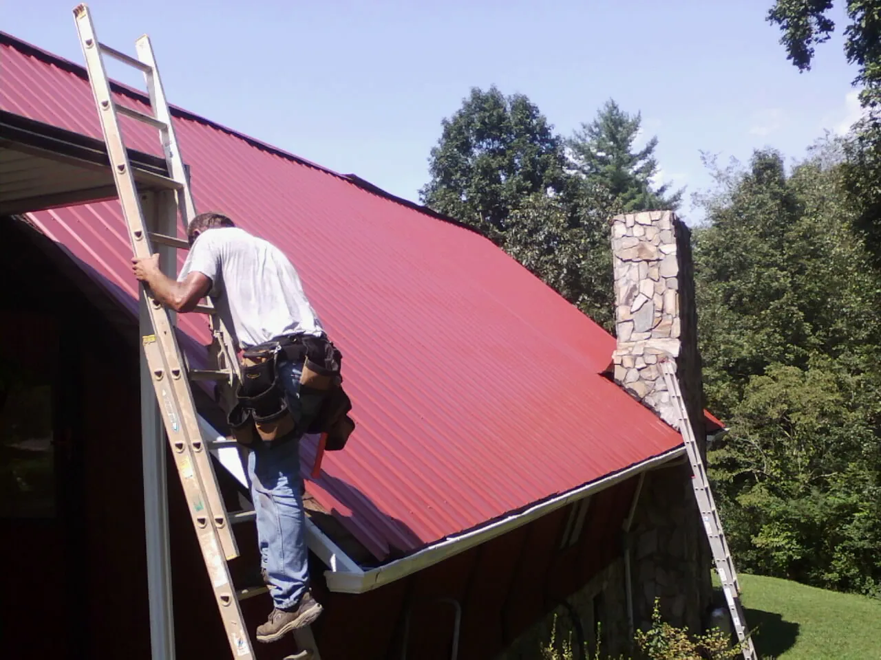 Roofer on a ladder beside a red metal roof and stone chimney — Soffit & Fascia Repair by Roofing Brothers in Tiffin